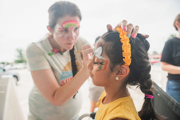Girl with rainbow on forehead giving another girl face paint