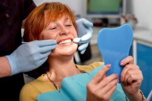 Woman with dental implants smiling into mirror at dentist's office.