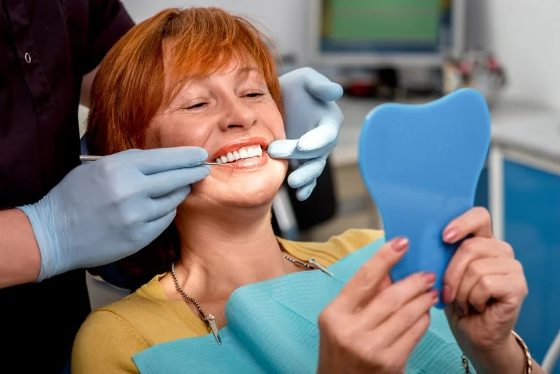 Woman with dental implants smiling into mirror at dentist's office. 